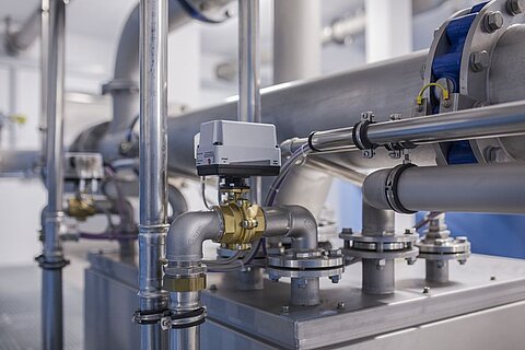 Eine Detailaufnahme von industriellen Rohrleitungen und Armaturen, einschließlich Ventilen und Sensoren. Die Rohre sind aus Metall und verleihen der Umgebung eine moderne, technische Atmosphäre. Der Hintergrund zeigt eine helle, saubere Industrieumgebung.