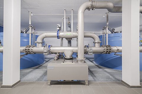 Industrieanlage mit großen blauen Wassertanks und einem Netzwerk aus Rohrleitungen. Sichtbare Ventile und Komponenten aus Metall verbinden die Rohre. Der Boden ist gefliest, und die Wände sind neutral gehalten.