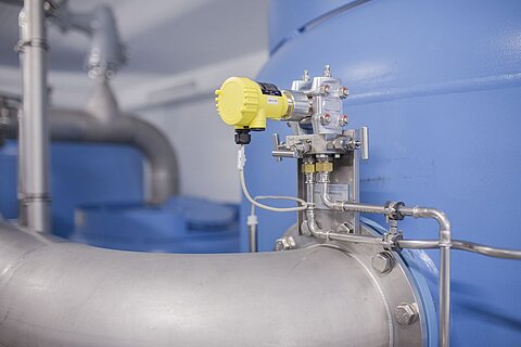 Bild eines Industrieanlagenbereichs mit blauen Tanks im Hintergrund. Im Vordergrund sind Rohrleitungen und ein Messgerät zu sehen, das zur Überwachung von Druck oder Durchfluss verwendet wird.