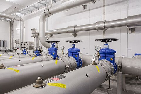 In einer Industriek Umgebung sind mehrere graue Rohre zu sehen, die mit blauen Ventilen ausgestattet sind. Einige der Ventile haben Manometer zur Druckmessung. Die Wände sind weiß, und es sind auch Metallleitungen sichtbar.