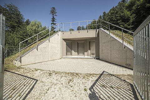 Ein Eingang zu einem unterirdischen Bunker, umgeben von Grünanlagen. Der Weg führt zu einer gepflasterten Fläche mit Geländer und zwei geschlossenen Türen. Im Hintergrund stehen hohe Bäume unter blauem Himmel.