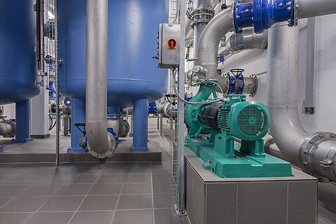 Das Bild zeigt eine industrielle Anlage mit blauen Wassertanks und einer grünen Pumpe. Rohrleitungen führen zu und von den Tanks, und eine Steuerbox ist an der Wand angebracht. Der Boden ist mit grauen Fliesen ausgelegt, und die Umgebung wirkt sauber und gut organisiert.