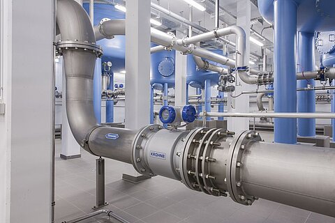 Innenansicht einer modernen Wasseraufbereitungsanlage mit großen blauen Speichertanks und metallischen Rohrleitungen. Die Rohre sind überwiegend aus rostfreiem Stahl und verlaufen durch den Raum, wobei einige Ventile und Messinstrumente sichtbar sind.