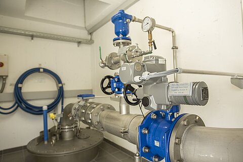 Ein Pumpensystem in einem Technikraum mit blauen und grauen Ventilen, Druckanzeige und Rohren. Im Hintergrund sind Wasserschläuche sichtbar.