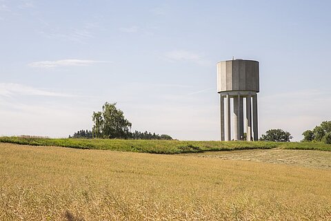 Ein Wasserturm steht auf einer grünen Wiese, umgeben von einem Feld mit reifem Getreide. Der Himmel ist klar und blau, mit wenigen Wolken. Im Hintergrund sind Bäume und eine sanfte Hügellandschaft zu sehen.