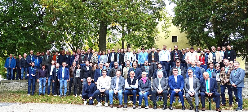 Gruppenfoto von etwa 100 Personen, die im Freien stehen und sitzen. Sie sind in verschiedenen Kleidungsstücken, darunter Hemden, Jacken und Anzüge, gekleidet. Im Hintergrund sind Bäume und ein Gebäude sichtbar. Die Menschen lächeln und scheinen an einem Treffen oder einer Veranstaltung teilzunehmen.