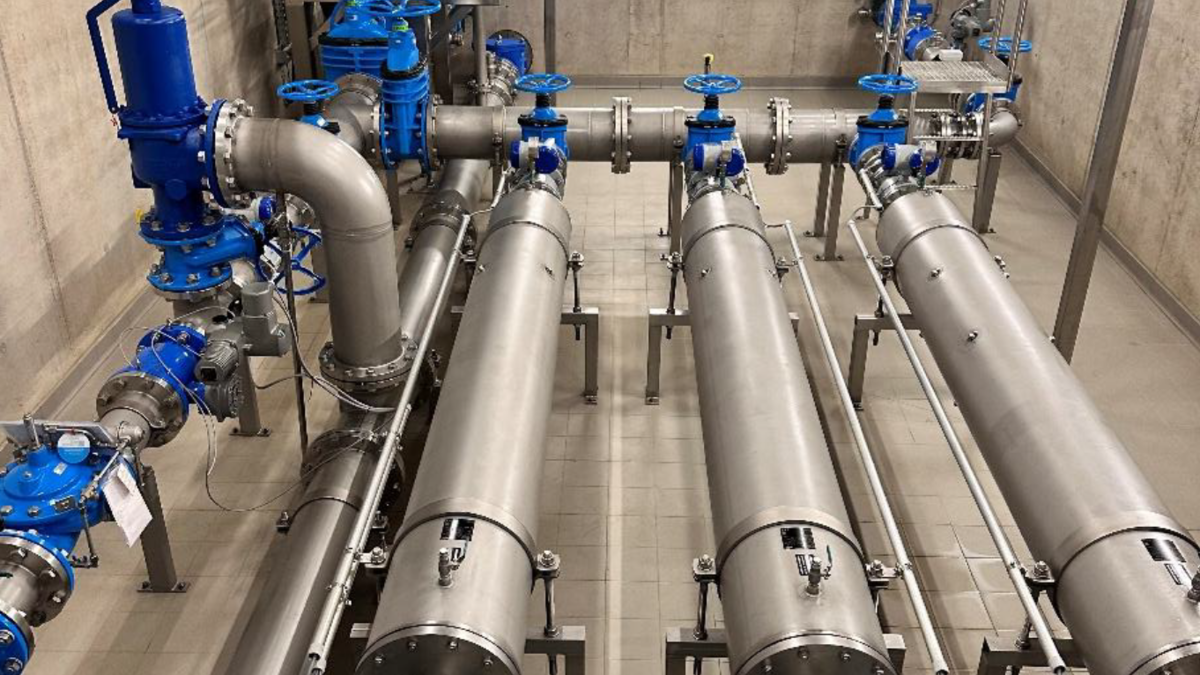 Eine industrielle Anlage mit Rohren, Ventilen und Pumpen. Die meisten Rohre sind aus Edelstahl, einige Teile sind blau. Der Boden besteht aus Fliesen und die Wände sind aus Beton. Die gesamte Einrichtung sieht modern und gut strukturiert aus.
