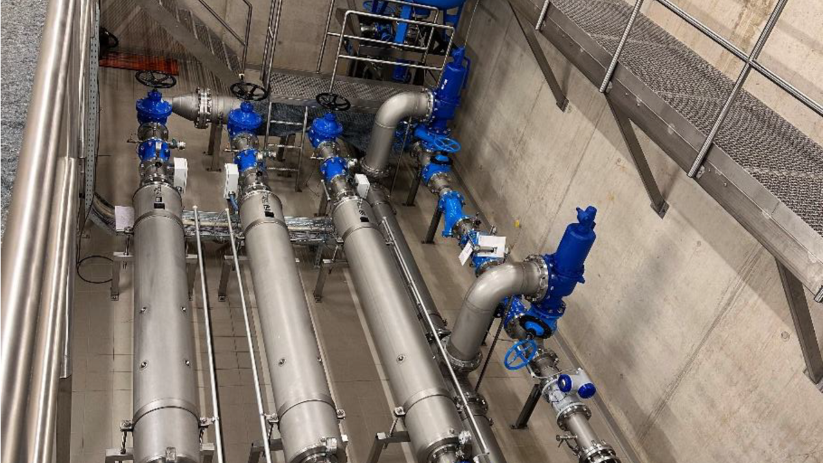 Ein Blick auf eine industrielle Rohrleitung, die aus mehreren großen silbernen Rohren und blauen Armaturen besteht. Die Rohre sind sauber angeordnet, und es gibt eine angelegte Metallplattform im oberen Bereich. Die Wände sind aus Beton und die Umgebung ist gut beleuchtet.