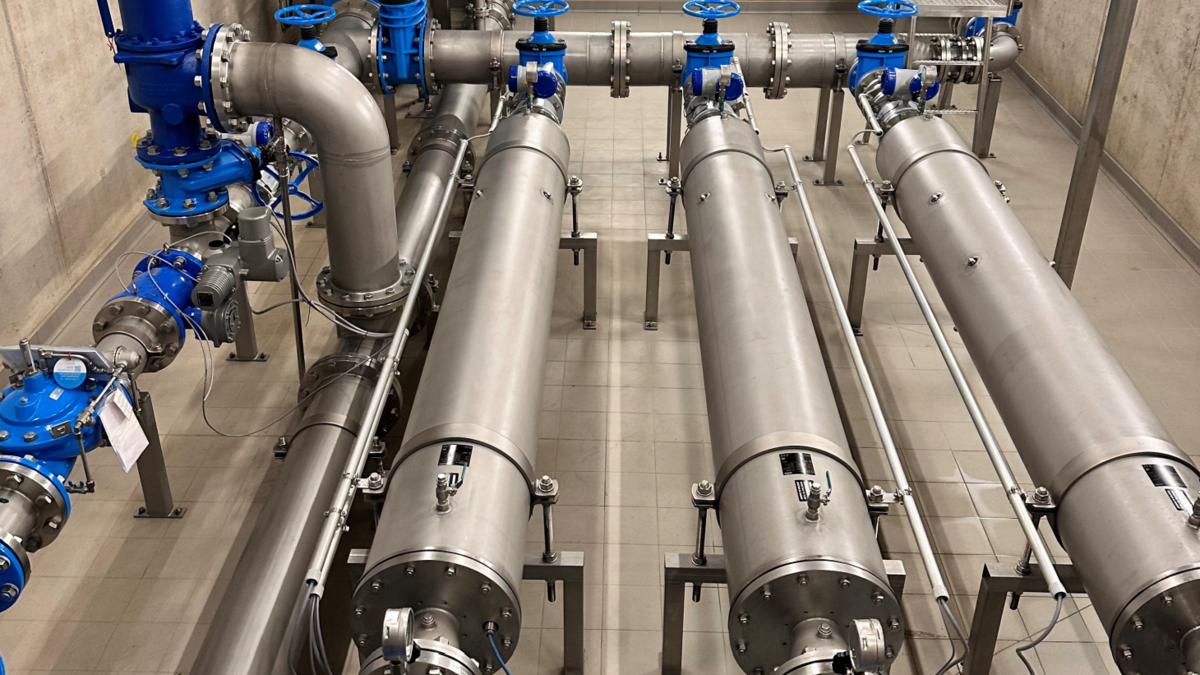 Ein technischer Raum mit mehreren langen, silberfarbenen Rohren, die auf Metallgestellen angeordnet sind. An den Rohren sind blaue Ventile und Kupplungen zu sehen. Der Boden ist gefliest und die Wände sind aus Beton.