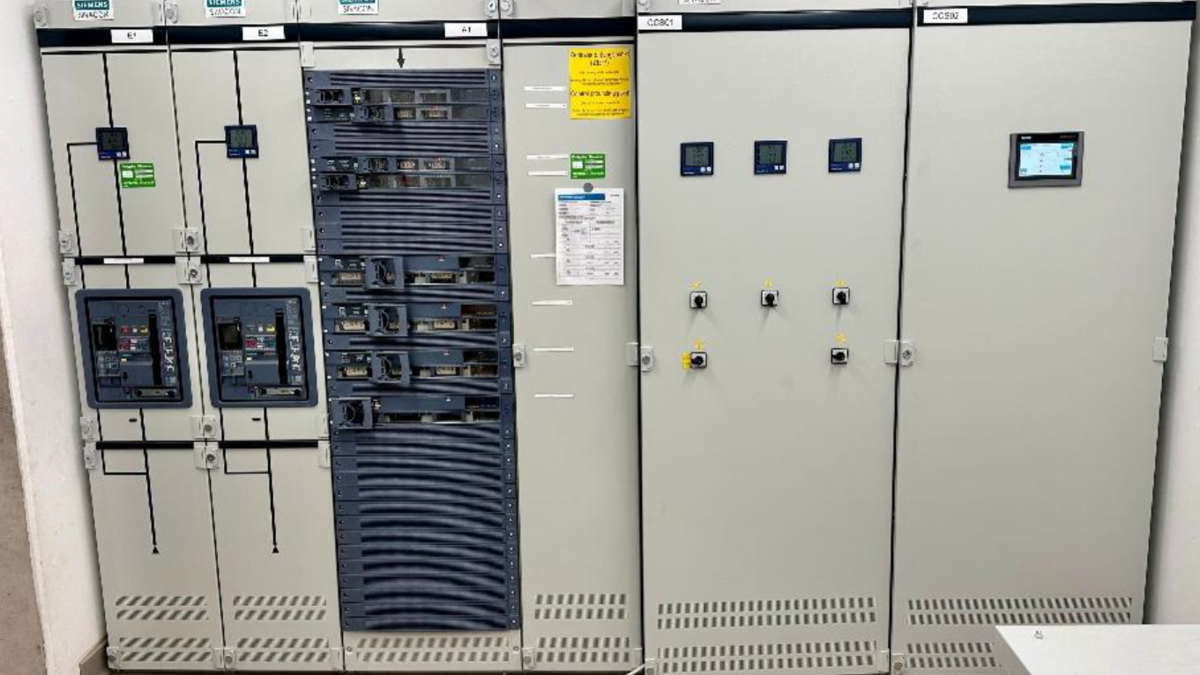 Ein Schaltschrank mit mehreren Steuergeräten und Schaltern. Auf der linken Seite sind verschiedene elektrische Schaltkästen mit Bedienelementen, während die rechte Seite zusätzliche Steuer- und Anzeigeeinheiten zeigt. An der Wand sind Informationen und Hinweise angebracht.