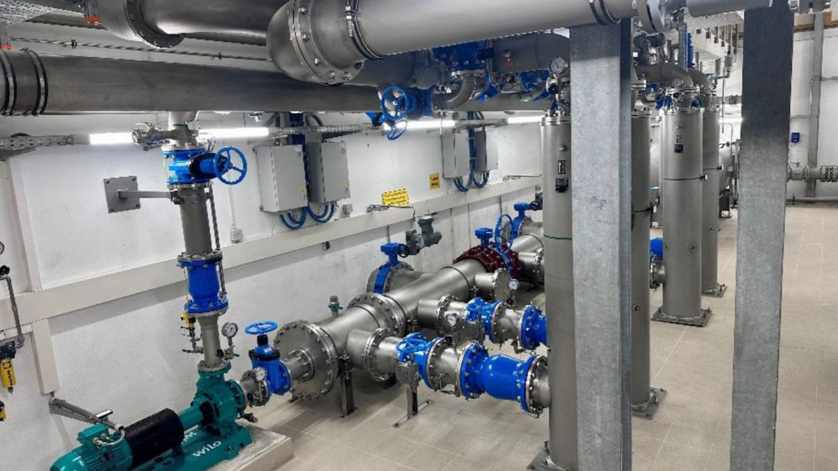 Moderne Rohrleitungsanlage mit silbernen und blauen Rohren, Ventilen und Pumpen. Verschiedene Apparateteile sind an der Wand montiert, während eine grüne Pumpe am Boden sichtbar ist. Der Raum hat helles Licht und einen sauberen, industriellen Look.