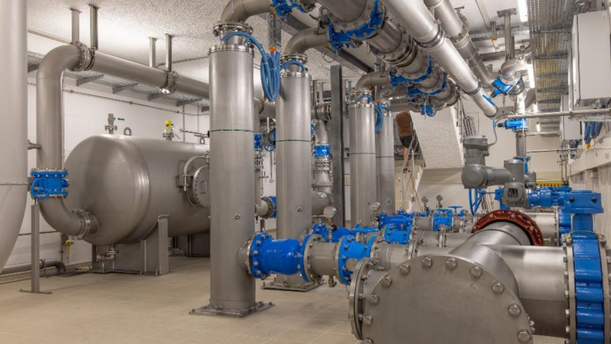 Eine industrielle Anlage mit großen silbernen Rohren, Ventilen und Maschinen. Ein Arbeiter steht auf einer Leiter, um an einer der Maschinen zu arbeiten. Die Rohre sind an verschiedenen Stellen blau markiert.