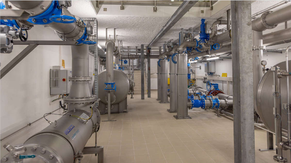 Das Bild zeigt eine industrielle Umgebung mit Rohren, Ventilen und Tanks, die in einem Prozessraum installiert sind. Die Wände sind hell, und die Böden sind gefliest. Alles wirkt sauber und gut organisiert, mit sichtbaren Anzeigegeräten und Steuerungen an den Wänden.