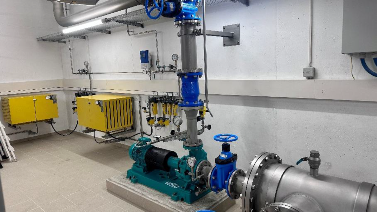 Ein Technikraum mit einer Pumpenanlage. Im Vordergrund ist eine grüne Pumpe mit einem Elektromotor und blauen Ventilen sichtbar. An der Wand sind gelbe Schaltschränke und verschiedene Druckmessgeräte angebracht. Die Wände sind aus Beton und der Boden ist gefliest.