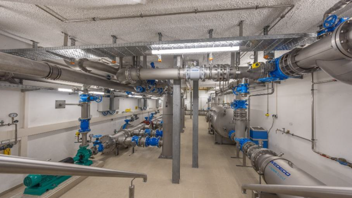 Ein Innenraum mit verschiedenen Rohren und Ventilen, die für die Wasseraufbereitung oder -verteilung verwendet werden. Metallische Oberflächen und blaue Komponenten sind sichtbar, sowie Pumpen und Messgeräte entlang der Wände. Der Boden ist gefliest und der Raum ist gut beleuchtet.