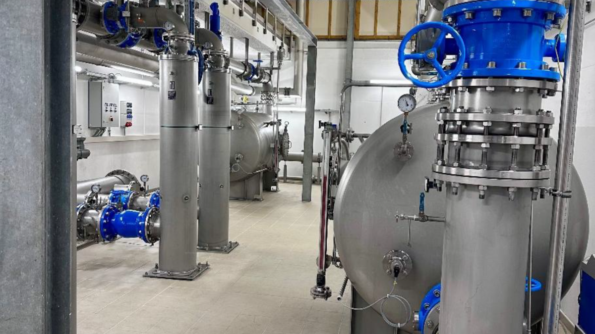 Der Innenraum einer industriellen Anlage mit mehreren großen, zylindrischen Hochbehältern aus Metall. Diese sind an Rohrleitungen angeschlossen, die blaue und silberne Elemente aufweisen. Die Wände sind hell und der Boden gefliest, mit verschiedenen Steuergeräten an einer Wand.