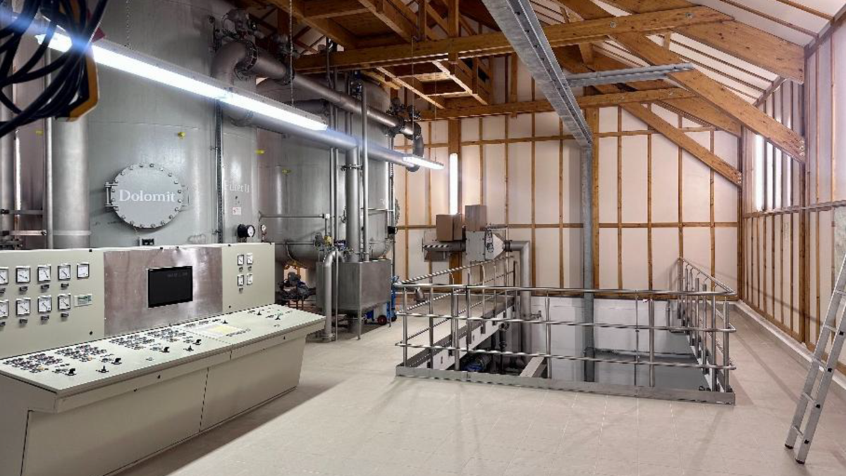 Ein industrieller Arbeitsraum mit großen silbernen Tanks und einem Steuerpult. Im Hintergrund sind Rohre und ein Geländer sichtbar, das zu einem tiefen Bereich führt. Der Raum hat Holzbalken an der Decke und eine helle Atmosphäre.