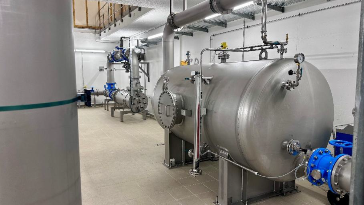 In einem industriellen Raum stehen mehrere große, kegelförmige Stahlbehälter. Leitungen und Ventile sind sichtbar, die eine komplexe Anlagentechnik darstellen. Der Boden ist gefliest und die Wände sind hell gestrichen. Künstliche Beleuchtung sorgt für eine helle Arbeitsumgebung.
