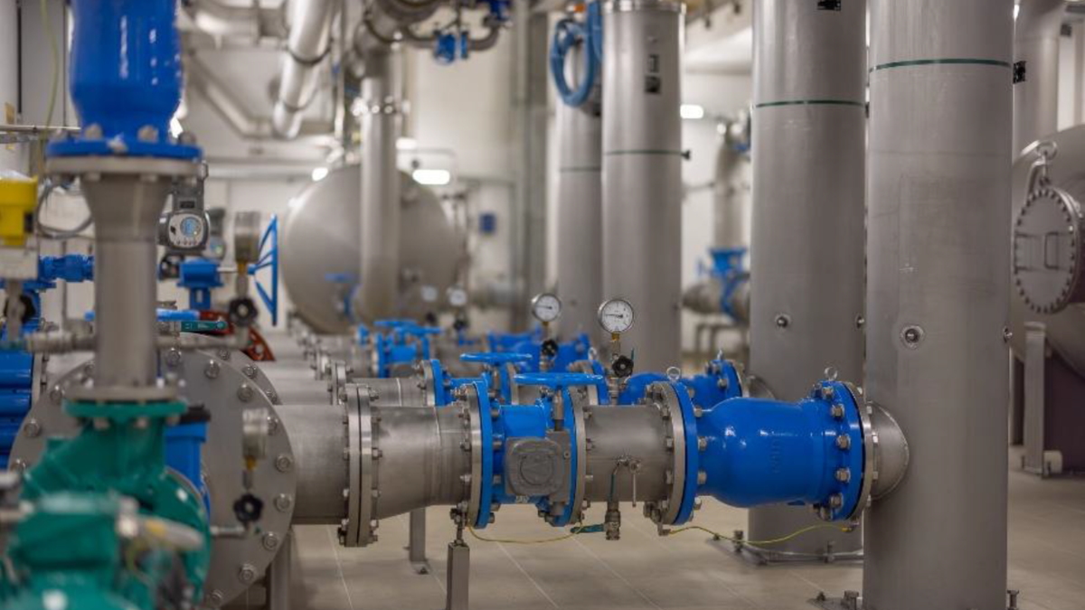 Industrieanlage mit Rohren und Ventilen in blauer und grauer Farbe. Mehrere Druckmesser sind sichtbar. Die Umgebung wirkt sauber und gut organisiert, mit verschiedenen Rohrleitungen und Tanks im Hintergrund.