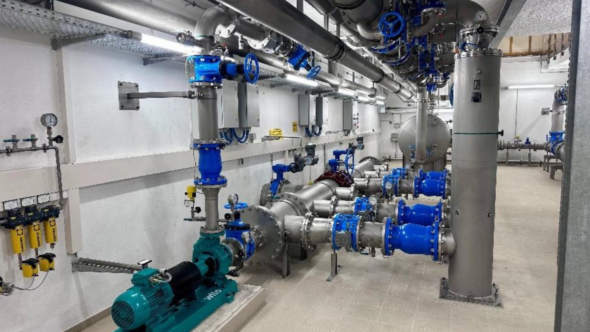 Eine technische Einrichtung mit Rohrleitungen und Ventilen in verschiedenen Größen. Sichtbare Pumpen und Messgeräte an der Wand. Der Boden ist gefliest. Die Einrichtung ist gut beleuchtet und in einem industriellen Stil gehalten.