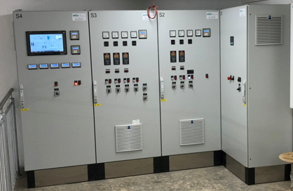 Eine Reihe von elektrischen Steuereinheiten mit verschiedenen Tasten, Schaltern und einem Bildschirm, die an einer Wand in einem Raum montiert sind. Die Einheiten sind in grauer Farbe und enthalten Edelstahl-Elemente am Boden.