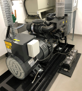 Eine Generatoranlage befindet sich in einem Raum. Die Maschine hat einen großen Motor und Kühlsystem, auf einem schwarzen Gestell montiert. Im Hintergrund sind weitere technische Geräte und eine Wand zu sehen.