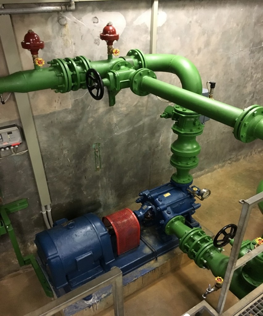 Vorher Ein Blick in einen Pumpenraum mit grünen Rohren und Ventilen. Eine blaue Pumpe mit einem roten Gitter befindet sich auf dem Boden. Die Wände sind aus Beton, und es gibt Kontrollpaneele an einer Wand. Der Raum ist beleuchtet und zeigt eine industrielle Umgebung.