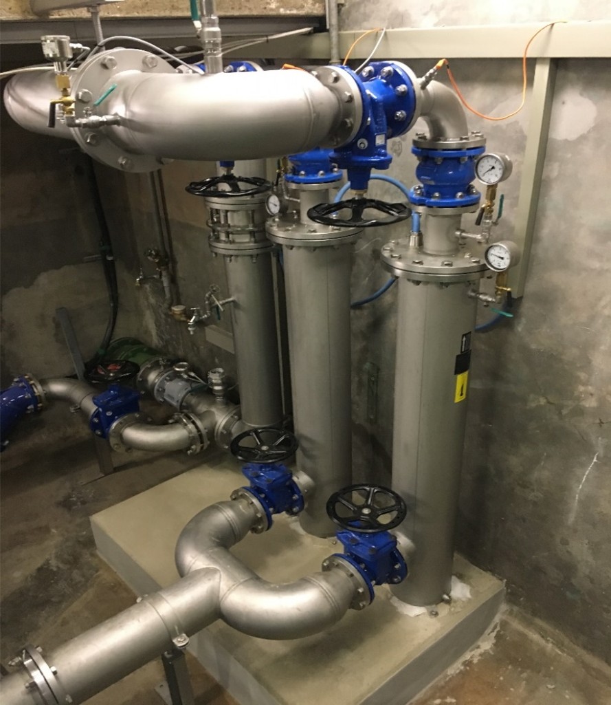 Nachher In einem Raum sind mehrere Metallrohre und Ventile installiert. Drei vertikale Rohre sind in Reihe angeordnet, mit blauen Ventilen und Druckmanometern. Eine gebogene Rohrleitung verbindet die Struktur, die auf einem betonsockel steht. Der Hintergrund ist aus grauem Mauerwerk.
