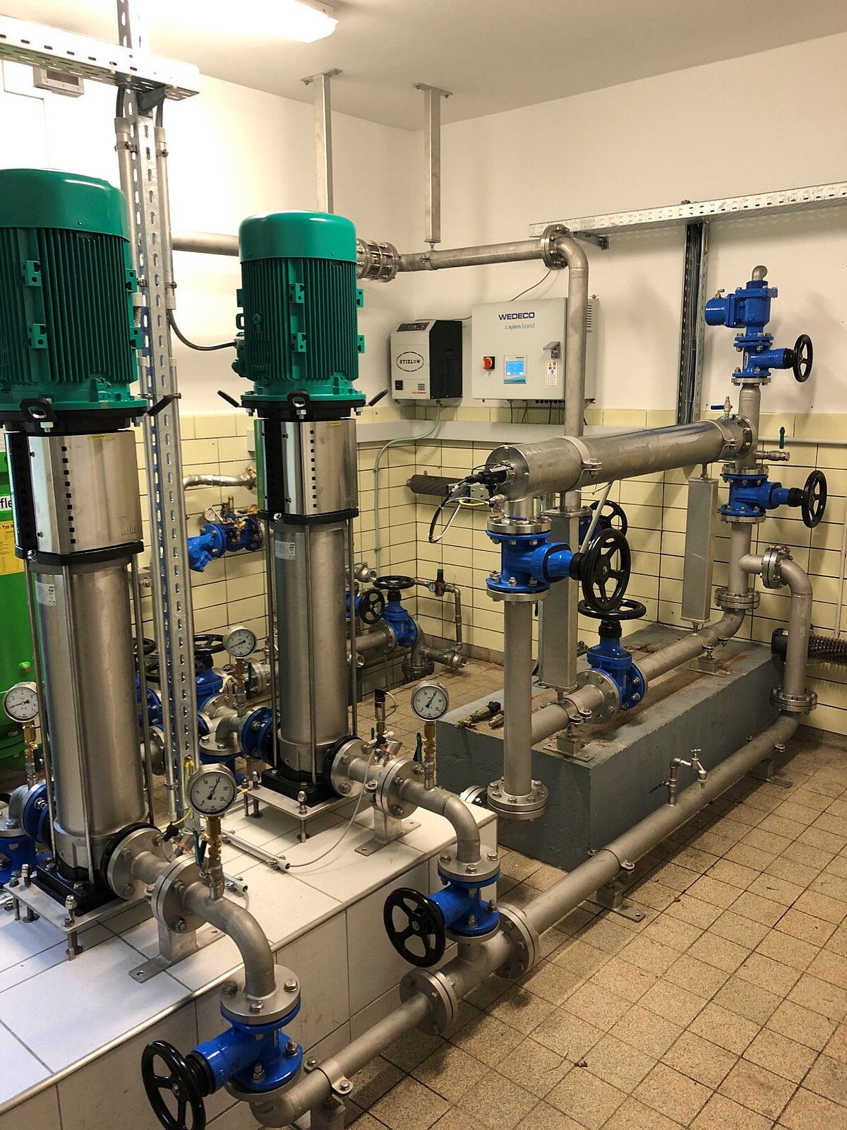 Eine technische Installation mit mehreren Pumpen, Rohren und Ventilen steht in einem Raum. Die Pumpen sind grün und aus Metall, während die Rohre und Ventile in Blau und Silber gehalten sind. Man sieht auch Druckmesser und eine Steuerungseinheit an der Wand.