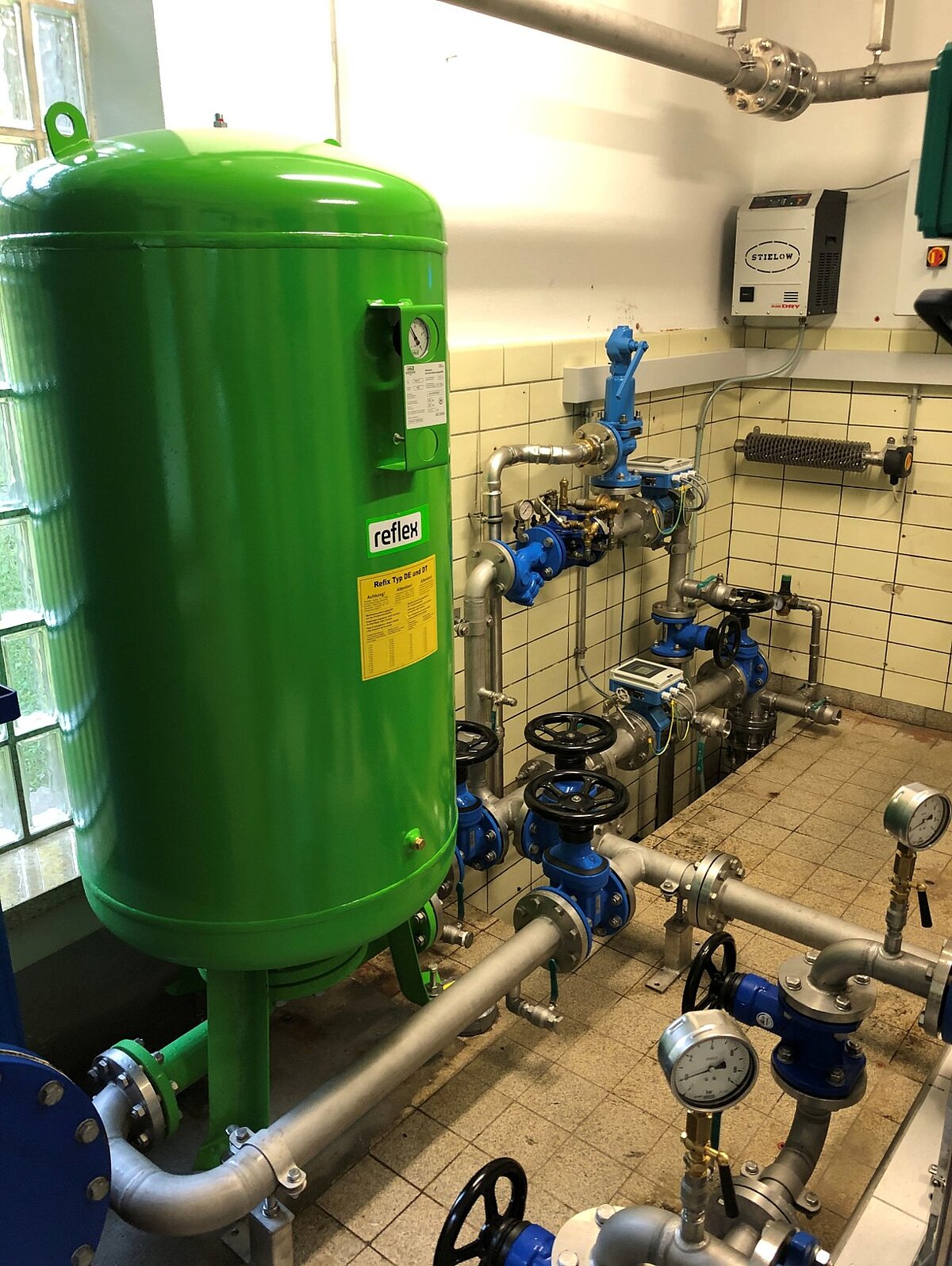 In einem technischen Raum sind Rohre und Armaturen sichtbar. Ein grüner Druckbehälter steht im Vordergrund, während blau gestrichene Ventile und Messgeräte im Hintergrund angeordnet sind. Die Wände sind gefliest und es sind einige Instrumente zur Überwachung installiert.