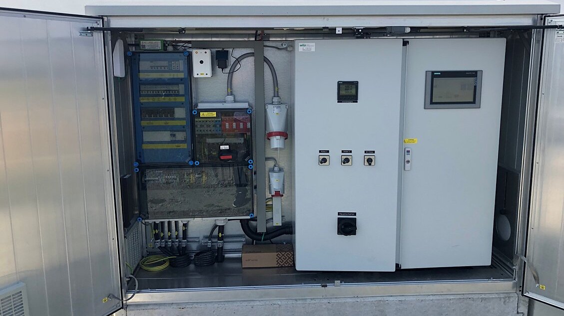 Ein geöffneter Schaltkasten mit verschiedenen elektrischen Komponenten, darunter Schalter, Kabel und ein Display. Das Innere ist strukturiert und zeigt eine Anordnung von Steuerelementen und Anschlüssen. Der Kasten steht an einem ruhigen Standort unter blauem Himmel.
