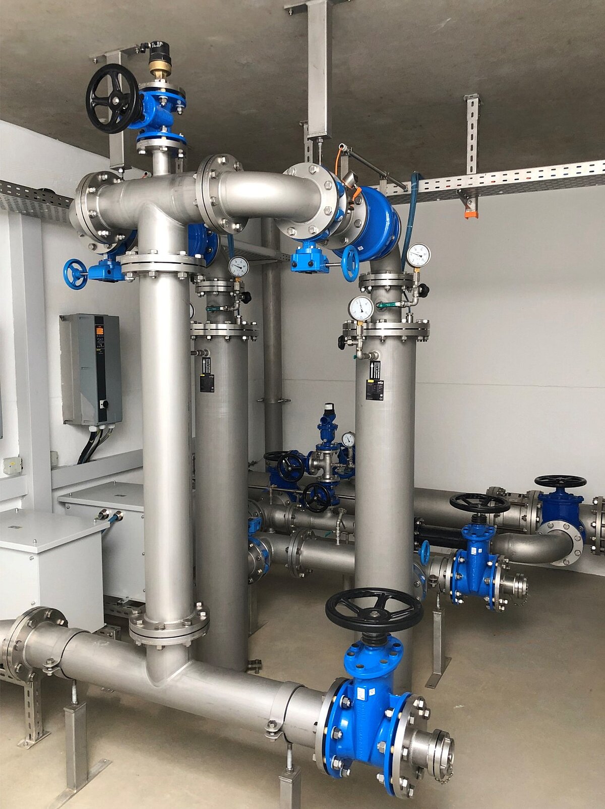 Eine industrielle Rohrleitungsanlage mit großen, silbernen Rohren und blauen Ventilen. Die Rohre verlaufen vertikal und horizontal, mit verschiedenen Ventiltypen und Druckmessgeräten. Die Umgebung ist schlicht und modern, mit einem Schaltschrank an der Wand.
