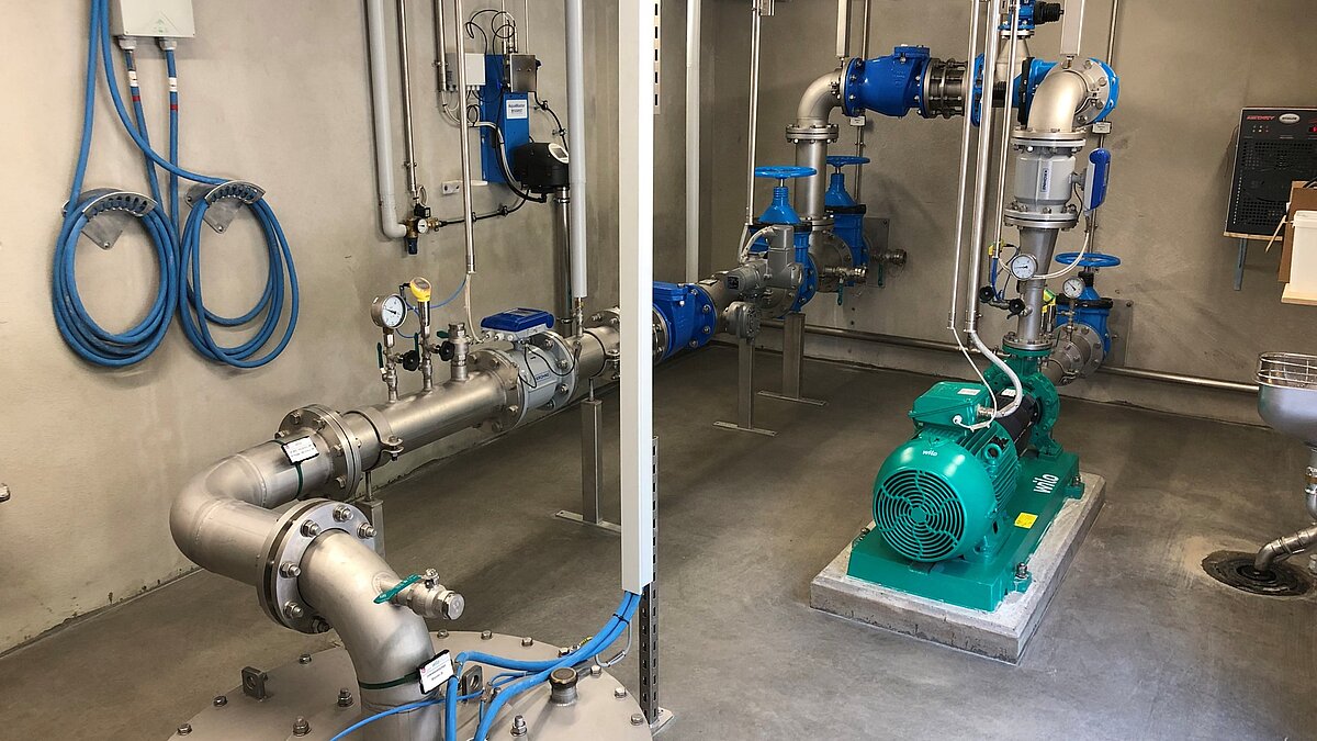 Eine industrielle Pumpstation mit verschiedenen Rohren und Ventilen. Rechts ist ein grüner Elektromotor sichtbar, während mehrere blaue Elemente und Messinstrumente auf den Rohren angebracht sind. Der Boden ist aus Beton, und die Wände sind grau.