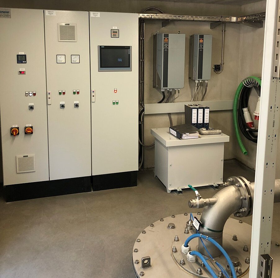 Ein technisch eingerichteter Raum mit elektrischen Steuerungspaneelen, einer Steuerkonsole und einem großen Rohrsystem. An der Wand sind mehrere elektronische Geräte montiert, und ein Tisch mit Dokumenten ist sichtbar. Beleuchtung erfolgt durch Deckenlampen.