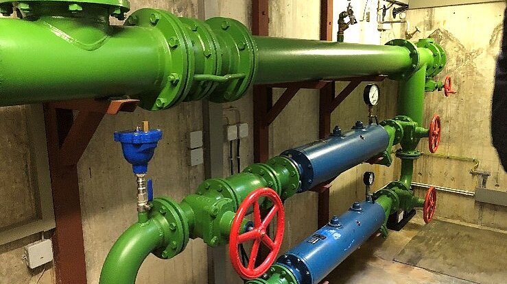 In einem Raum sind große Rohre zu sehen, hauptsächlich in Grün und Blau, mit roten Ventilen. An den Wänden sind einige Geräte und Messinstrumente angebracht. Die Rohre scheinen Teil eines technischen Systems zur Wasser- oder Flüssigkeitsverteilung zu sein.