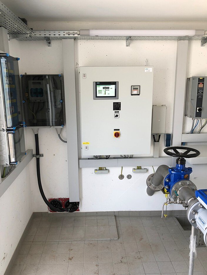 Ein Kontrollraum mit einem großen Steuergerät an der Wand, einem Display und mehreren Bedienelementen. Daneben befindet sich ein Ventil mit einem blauen Gehäuse. Die Wände sind schlicht und der Boden ist gefliest.