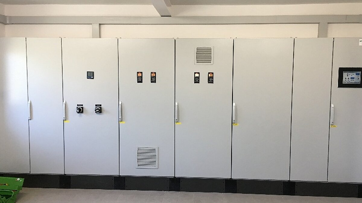 Eine Reihe von grauen elektrischen Schaltschränken an einer Wand, einige mit Kontrollknöpfen und Anzeigen. Die Schaltschränke sind geschlossen und stehen nebeneinander in einem gut beleuchteten Raum.