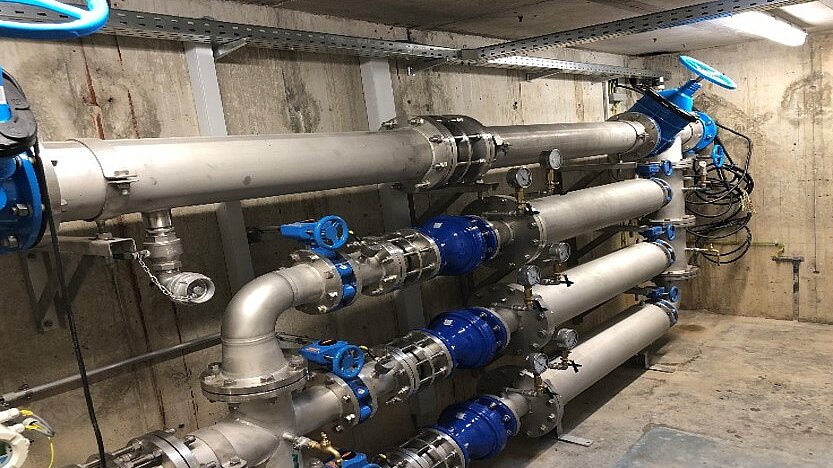 Im Bild sind mehrere große Rohre zu sehen, die an einer Wand montiert sind. Die Rohre bestehen aus Metall und haben blaue Armaturen. Sie sind Teil eines Rohrleitungssystems in einem technischen Umfeld, möglicherweise in einer Anlage zur Wasseraufbereitung.
