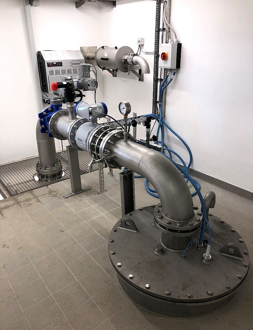 Ein industrielles Rohrleitungssystem mit einer großen Metallleitung, einem Ventil und verschiedenen Messgeräten. Im Hintergrund befindet sich eine Wand mit elektrischen Steuerungen. Der Boden ist gefliest, und die Umgebung sieht sauber und gut gewartet aus.