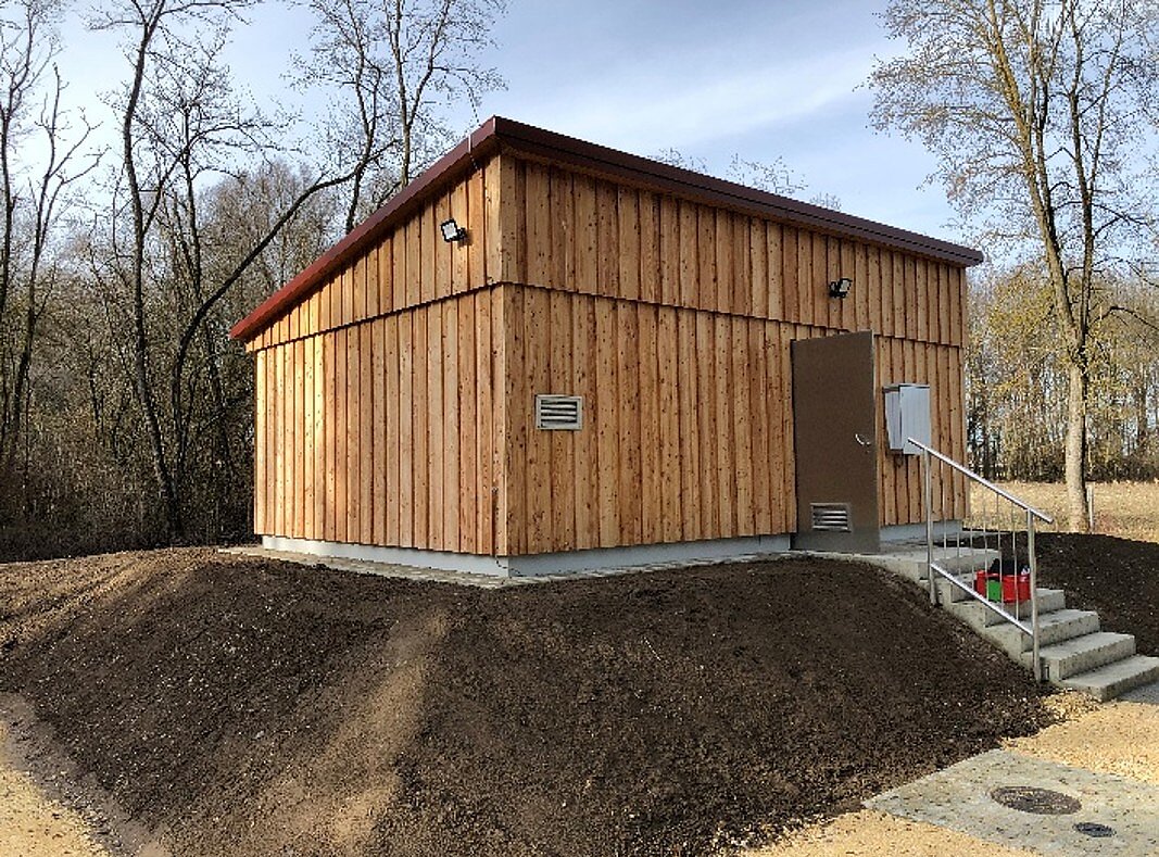 Ein modernes, aufgeständertes Holzgebäude mit einem geneigten Dach. Die Fassade ist aus Holzlatten und das Gebäude verfügt über ein paar Fenster sowie eine Eingangstür. Vor dem Gebäude befindet sich eine kleine Treppe und der Boden ist mit Erde aufgeschüttet. Bäume umgeben den Bereich.