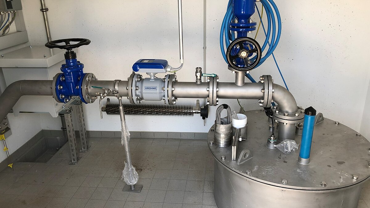 In einem Raum sind Rohre und Ventile sichtbar, die Teil einer technischen Installation sind. Ein blauer Regler und ein Metallbehälter sind ebenfalls zu sehen. Die Einrichtung scheint für die Kontrolle von Flüssigkeiten konzipiert zu sein.