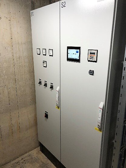 Zwei elektrische Steuerungsschränke in einem Betonraum. Der linke Schrank trägt die Bezeichnung "S1" mit verschiedenen Bedienknöpfen und Anzeigen. Der rechte Schrank, "S2", verfügt über ein Display und eine Handbedienung. Die Wände sind aus Beton und der Boden ist unbehandelt.