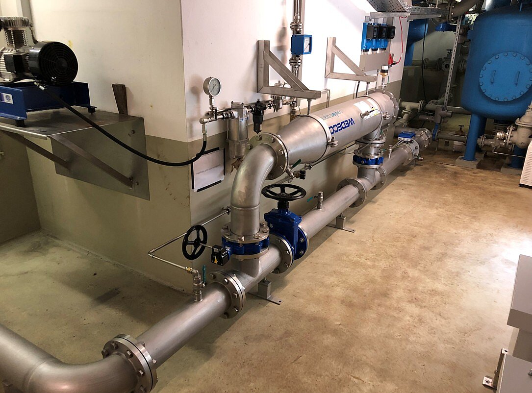 Ein Bild einer industriellen Rohrleitungsinstallation. Sichtbar sind große, metallene Rohre mit Ventilen und Manometern. Im Hintergrund befindet sich ein großes, blaues Gerät, das vermutlich eine Pumpe oder einen Wassertank darstellt. Der Boden hat einen einfachen, glatten Betonbelag.