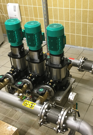 Das Bild zeigt eine Anlage mit drei Wassermotoren, die auf einem Montagerahmen installiert sind. Die Motoren haben grüne Abdeckungen und sind mit Metallrohren sowie Ventilen verbunden. Der Hintergrund besteht aus gefliesten Wänden in einem Technikraum.