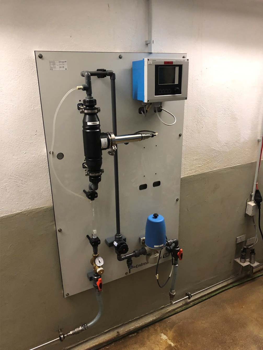 An einer Wand montiertes technisches Gerät mit verschiedenen Rohren und Ventilen. Es enthält einen Zähler, einen Druckregler und einen Kontrollmonitor. Die Installation scheint für die Steuerung eines Flüssigkeitsstroms konzipiert zu sein.